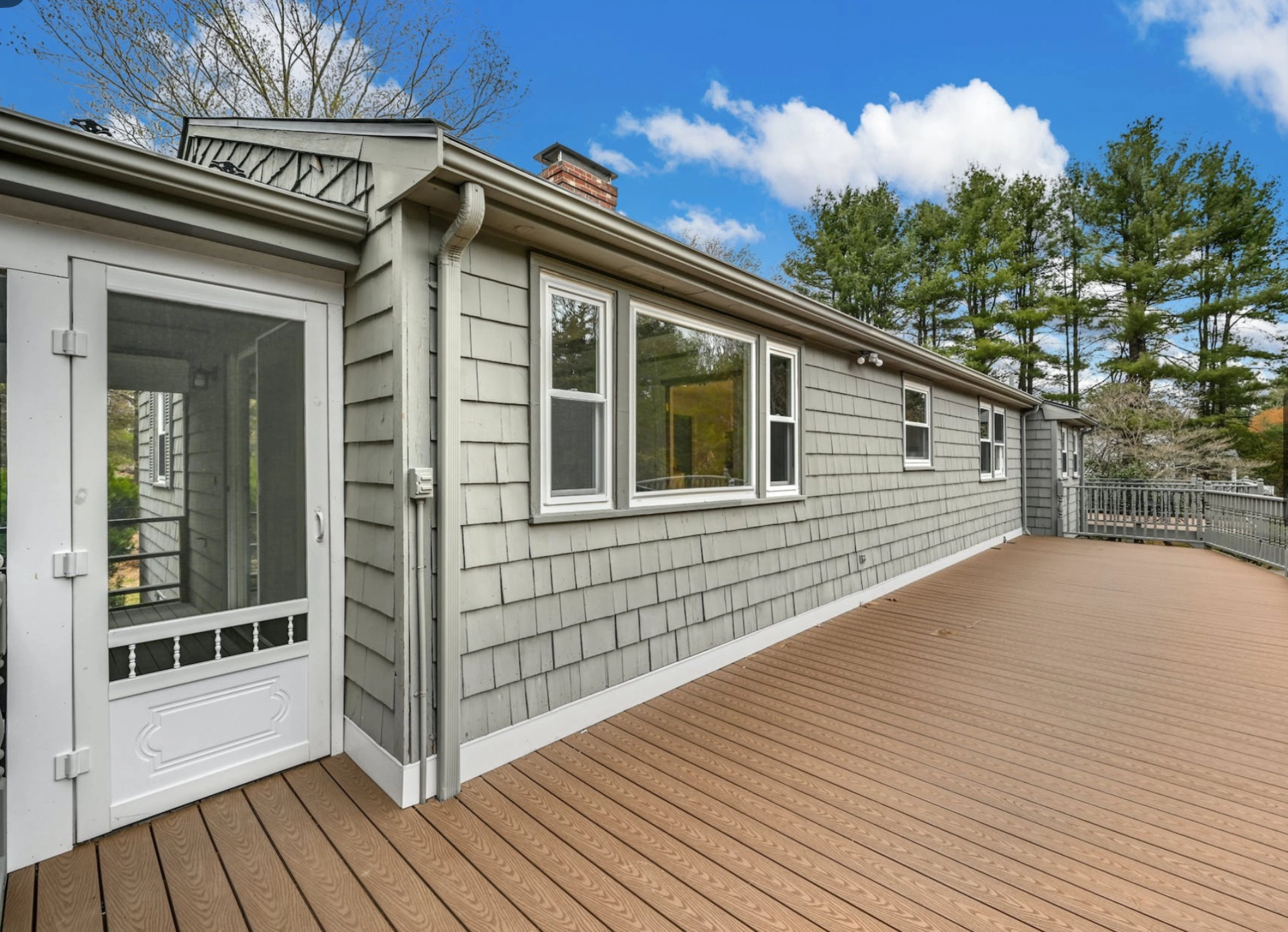 House with gray shingle siding, white framed windows, and a large wooden deck, bordered by trees under blue sky.