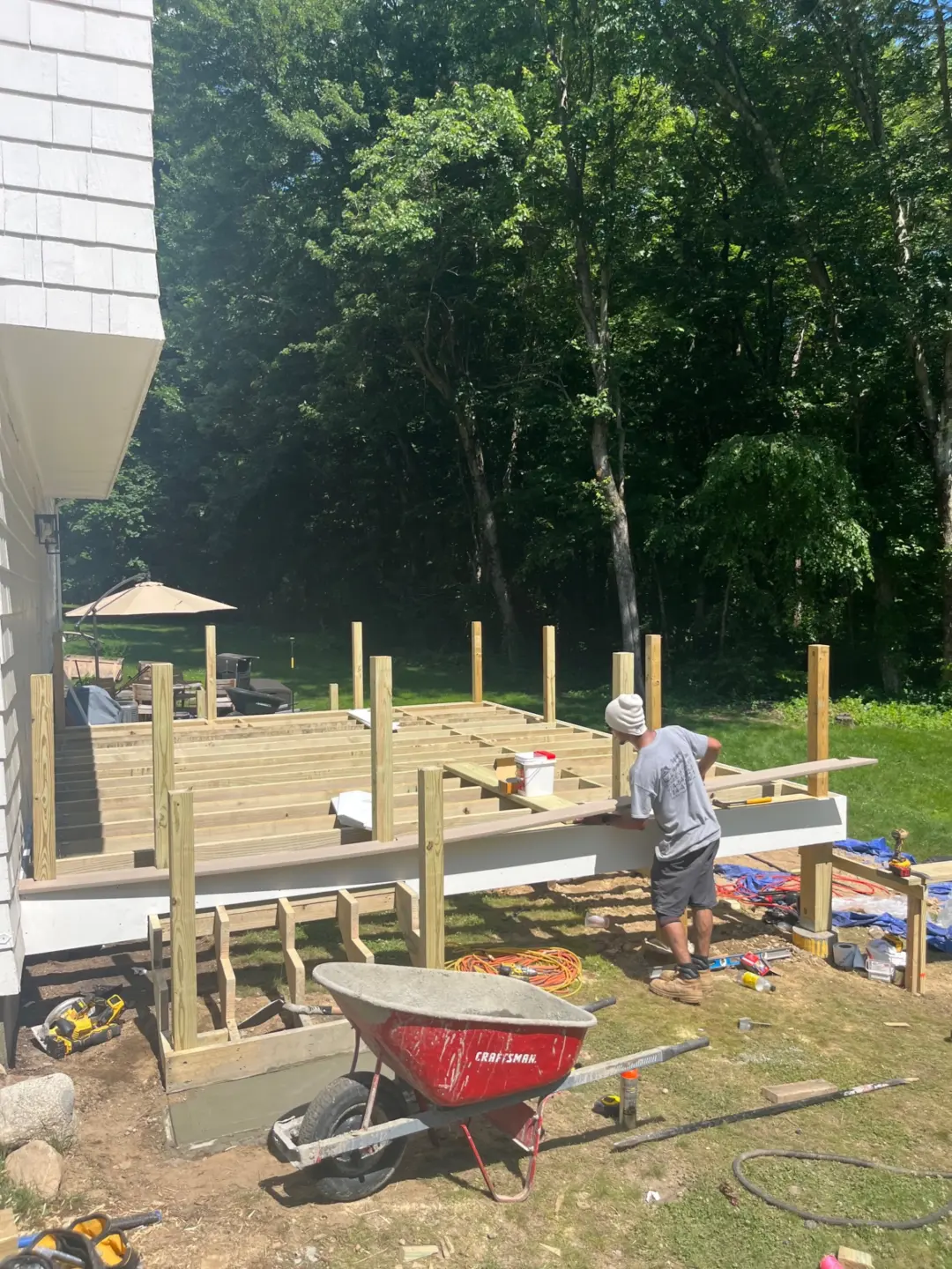 Worker constructing wooden deck beside house with tools and supplies scattered on grass, surrounded by trees.
