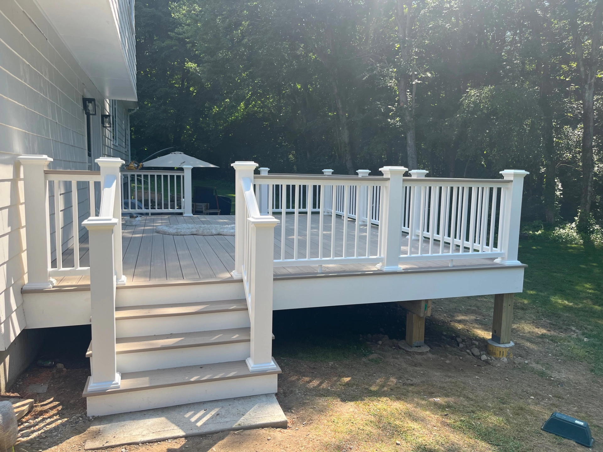 Wooden deck with white railing and stairs, adjacent to house, umbrella and chairs in background, surrounded by trees.