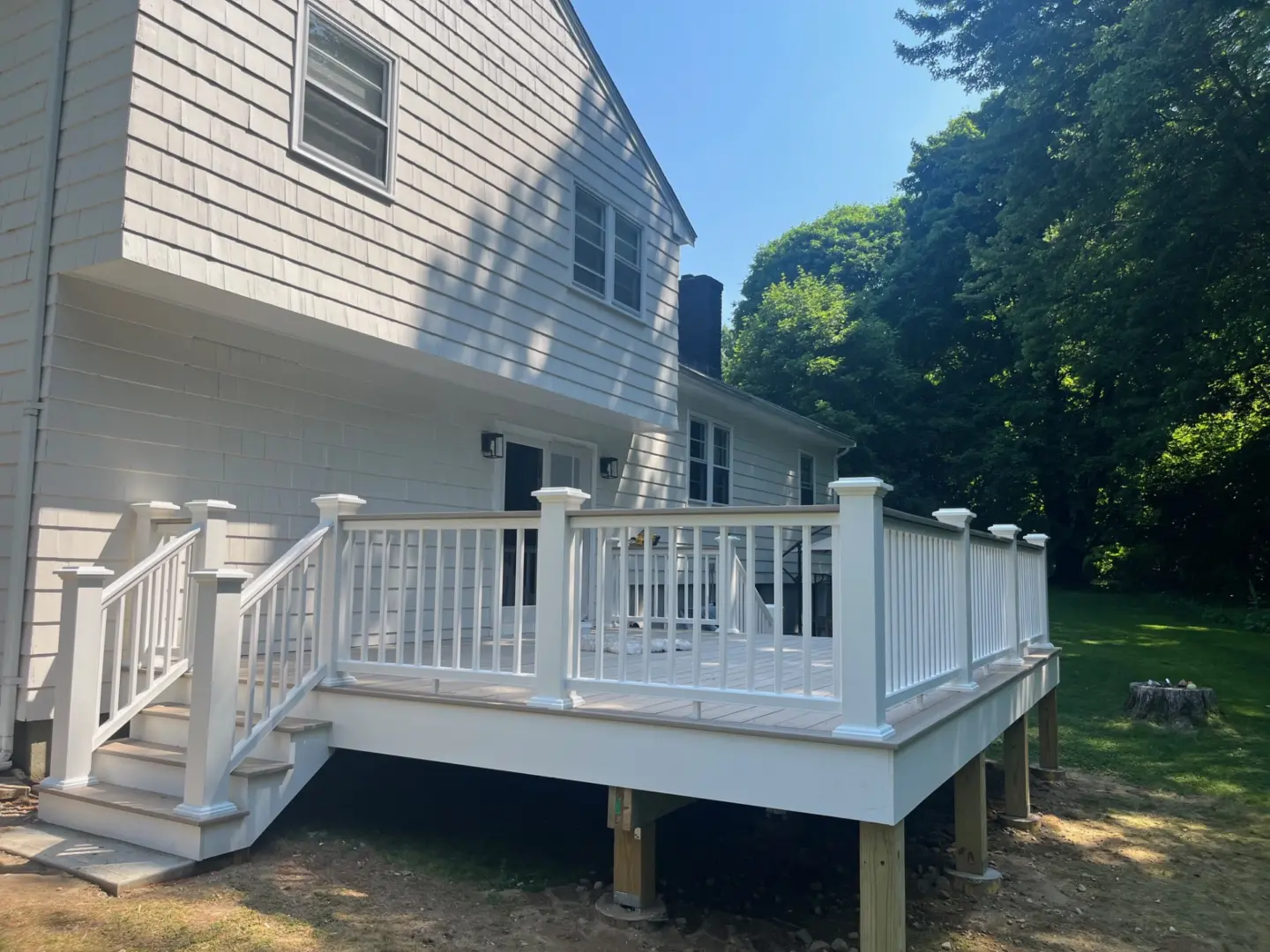White wooden deck with railing attached to a house, surrounded by grass and trees, with stairs leading down.
