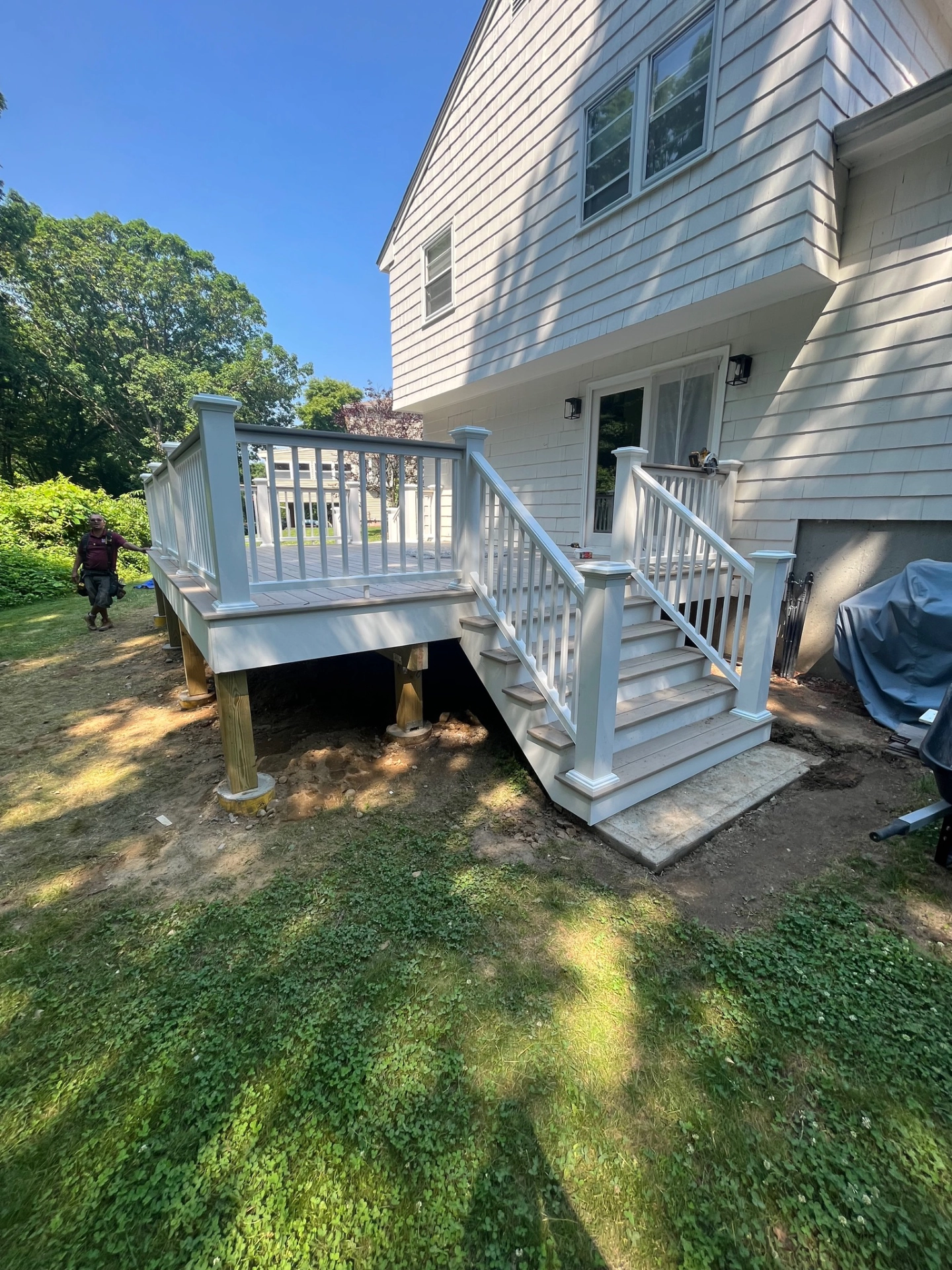 White deck with railing and steps attached to house, grass below, person walking on left, trees in background.