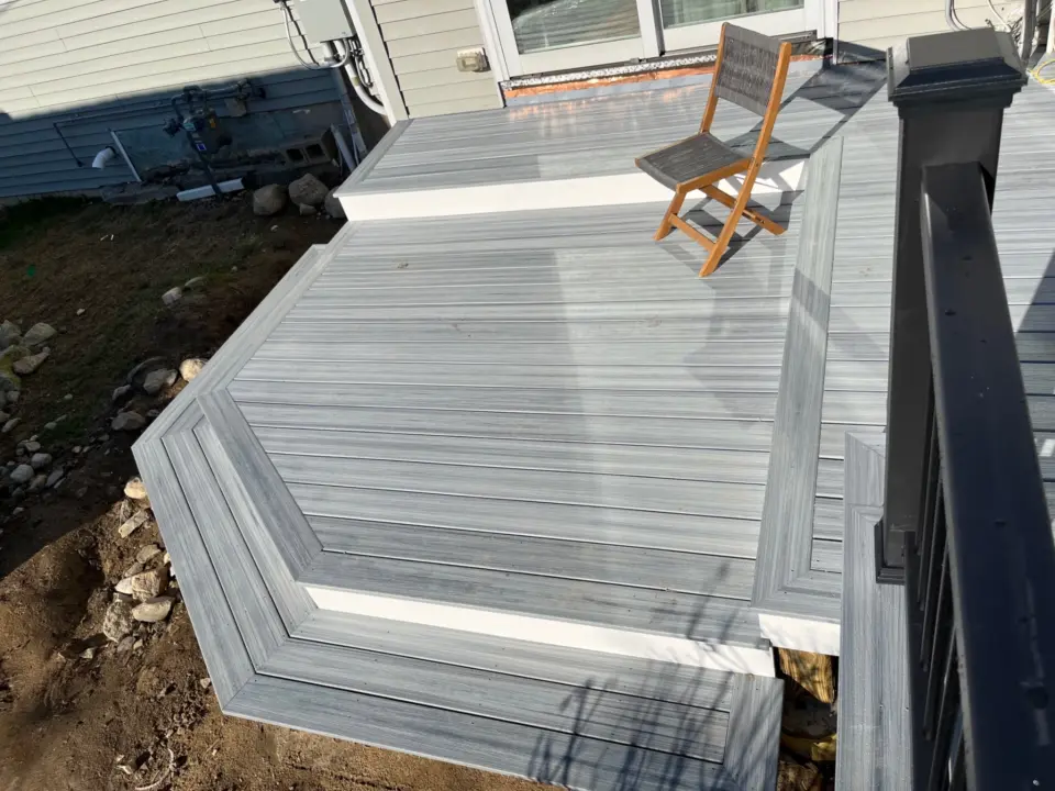Gray wooden deck with brown chair, adjacent to house, surrounded by dirt and stones, view of railing and siding.