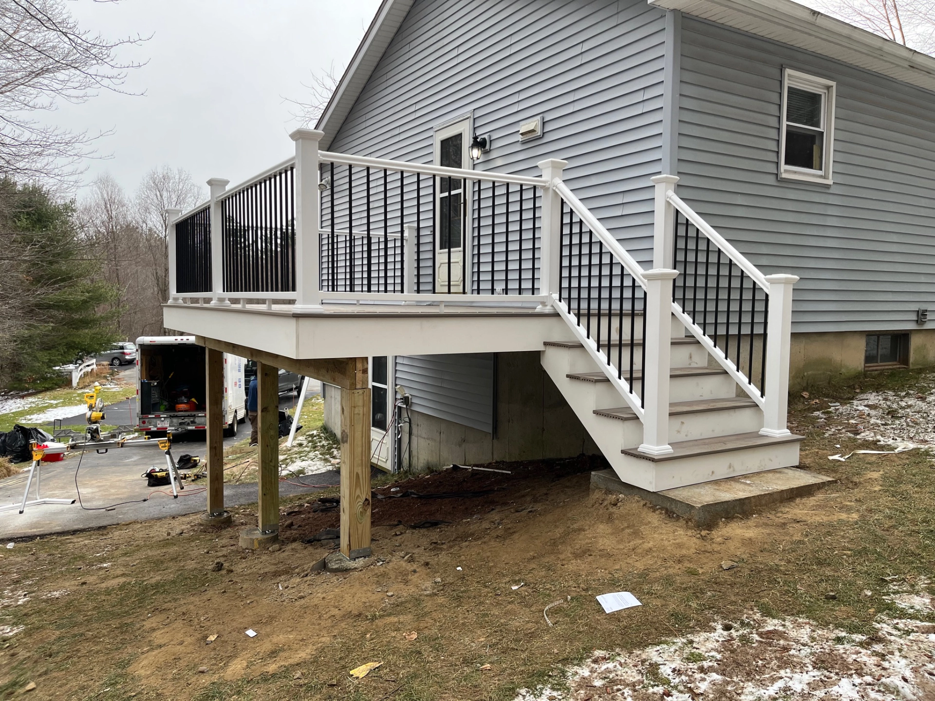 Elevated deck with white railing attached to grey house, stairs descending, construction tools and vehicle in background.