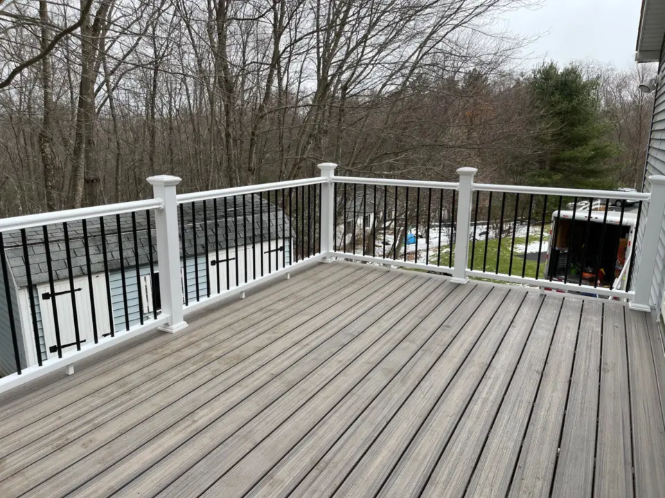 Wooden deck with white railing overlooking snow-dusted yard, trees, and parked trailer near a small shed.