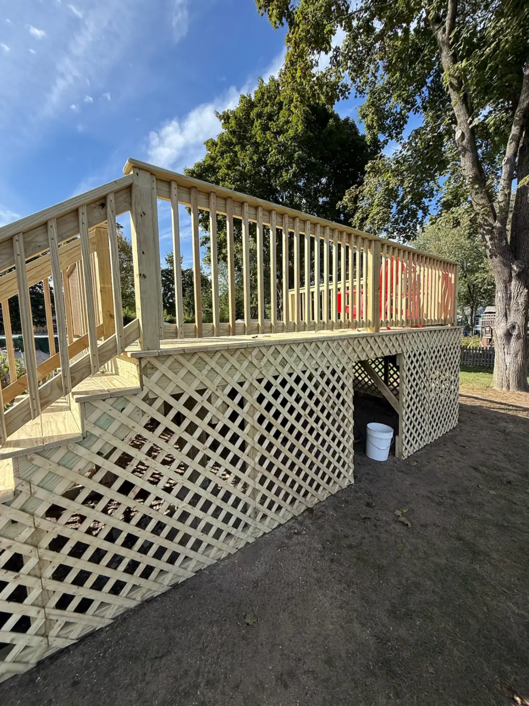 Wooden deck with lattice design, stairs leading down, surrounded by trees, white bucket beneath, blue sky overhead.