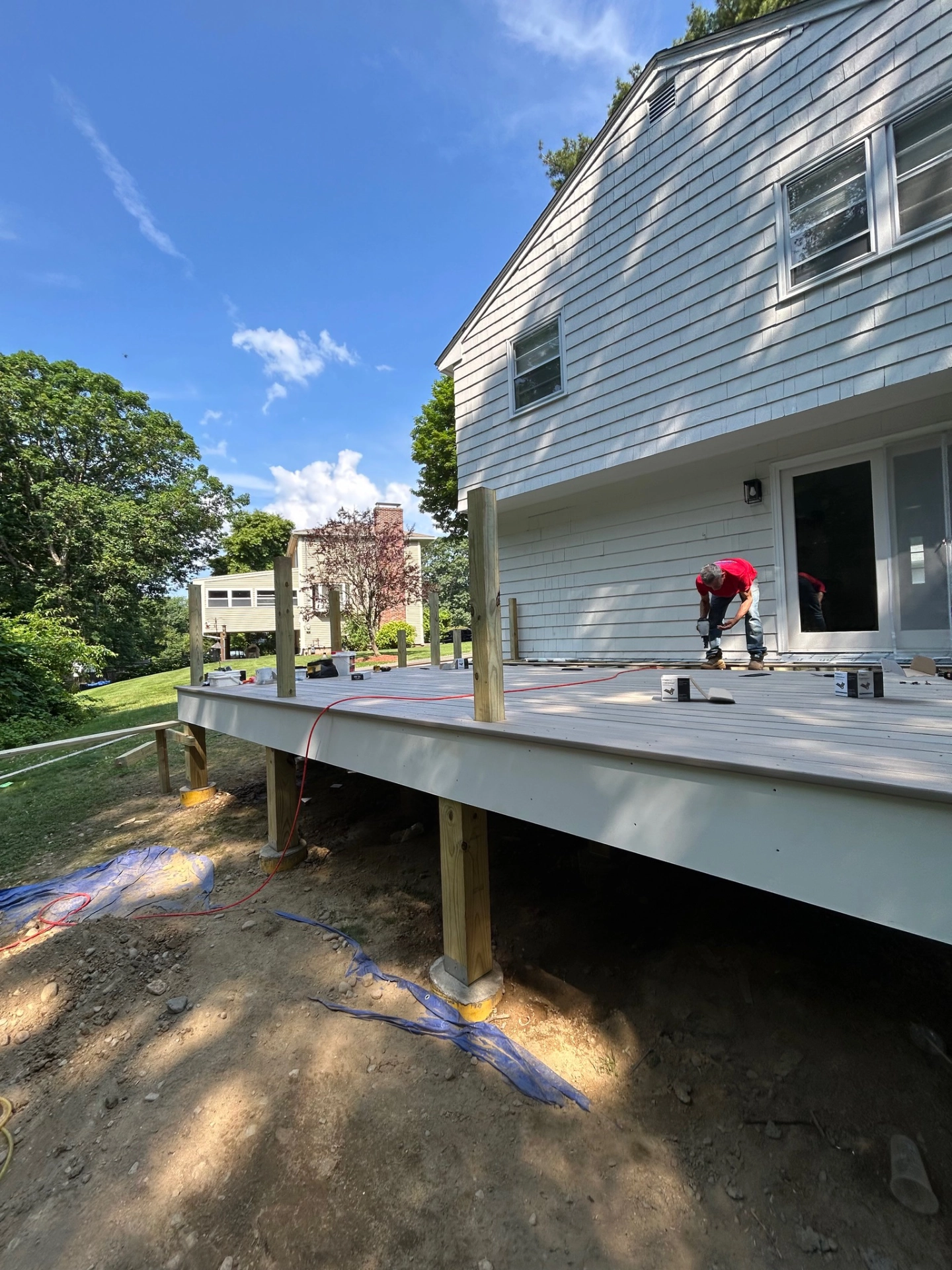 House exterior with new deck under construction, worker in red shirt on right, poles and tools visible, clear blue sky.