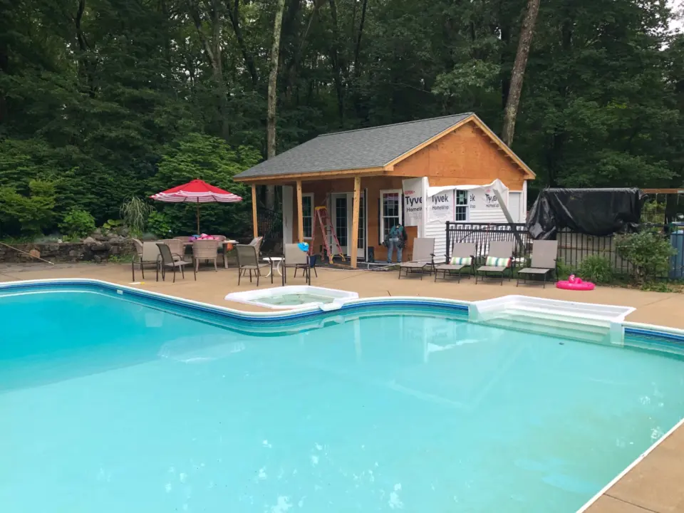 Swimming pool with patio chairs, red umbrella, construction in progress on building, surrounded by trees.