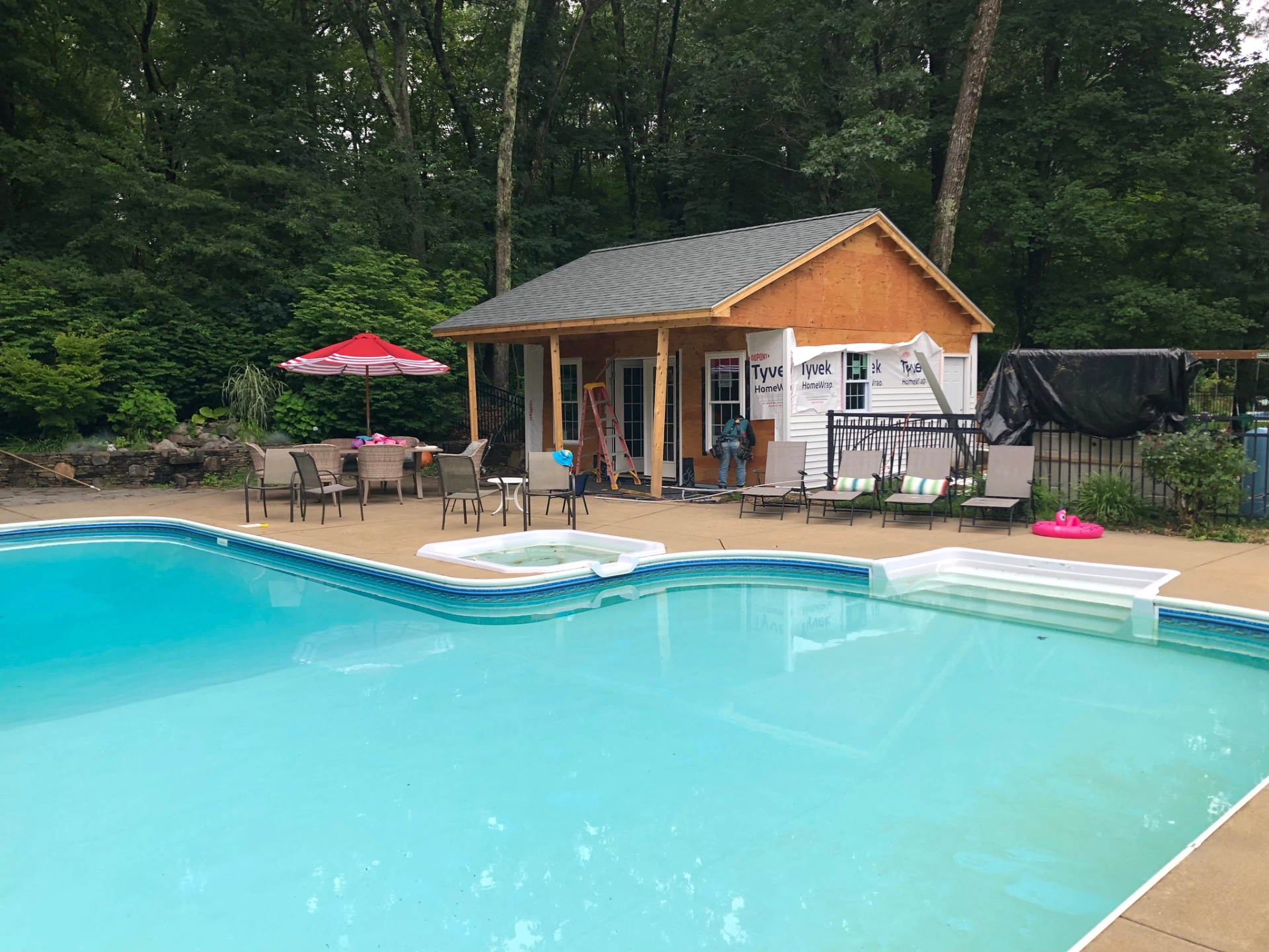 Swimming pool with patio chairs, red umbrella, construction in progress on building, surrounded by trees.