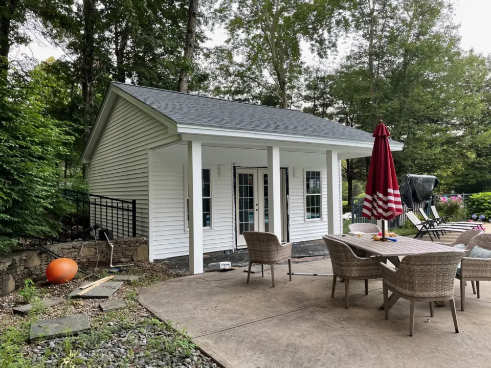 White shed with French doors beside a patio, red umbrella, wicker chairs around table, surrounded by trees.