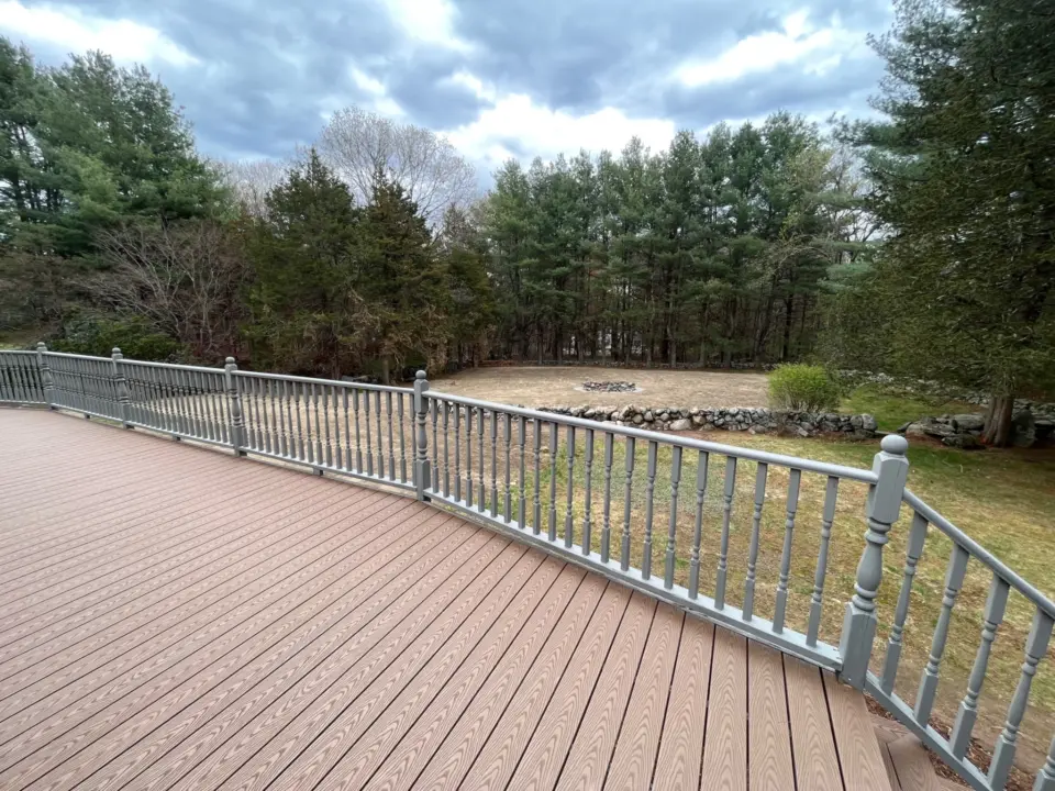 Wooden deck with gray railing overlooking grassy yard, stone firepit, and trees under a cloudy sky.