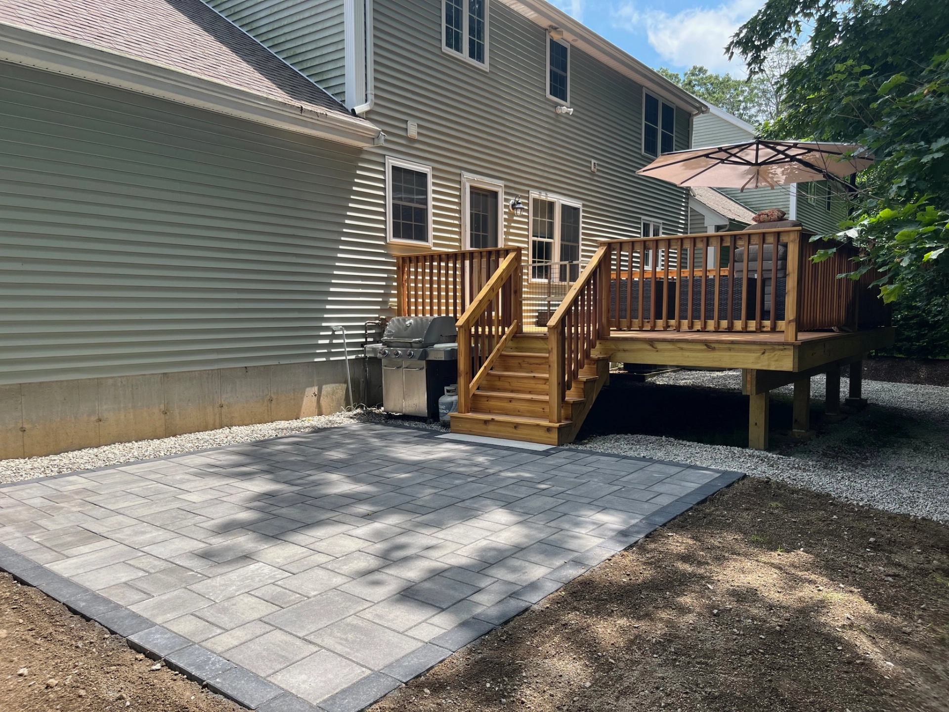 Wooden deck with stairs, umbrella, and grill beside house, gray stone patio in foreground, surrounded by trees.