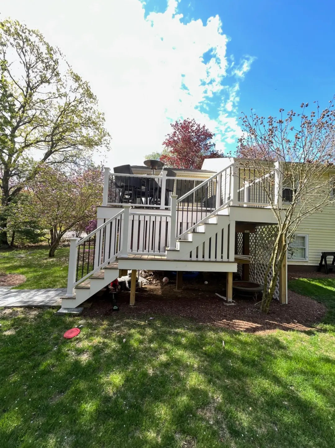Elevated wooden deck with stairs, outdoor furniture, and railing, surrounded by trees and grass under a blue sky.