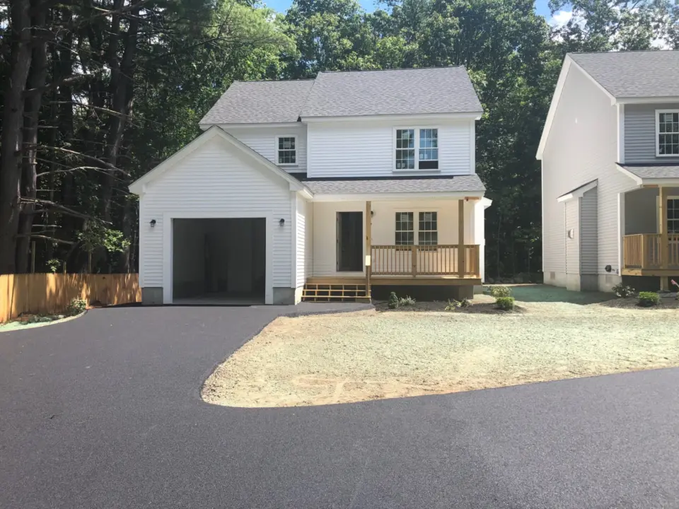 White two-story house with attached garage, front porch, black driveway, and wooden fence, surrounded by trees.