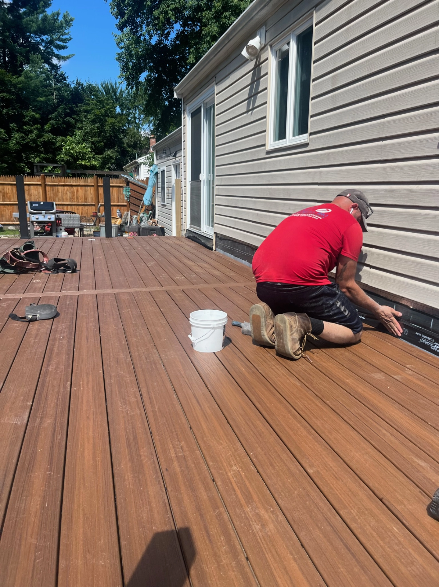 Person in red shirt installing trim on wood deck next to house, white bucket nearby, backyard visible in background.