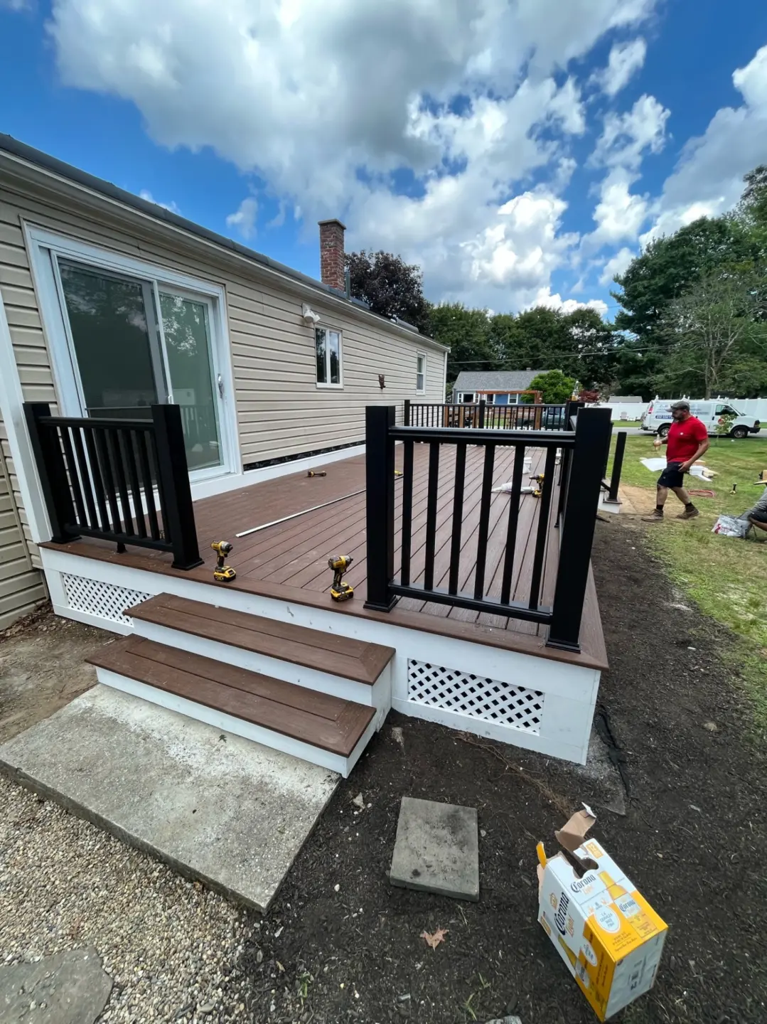 Wooden deck attached to beige house, black railings, power tools on deck, open lawn, person in red shirt with paper.