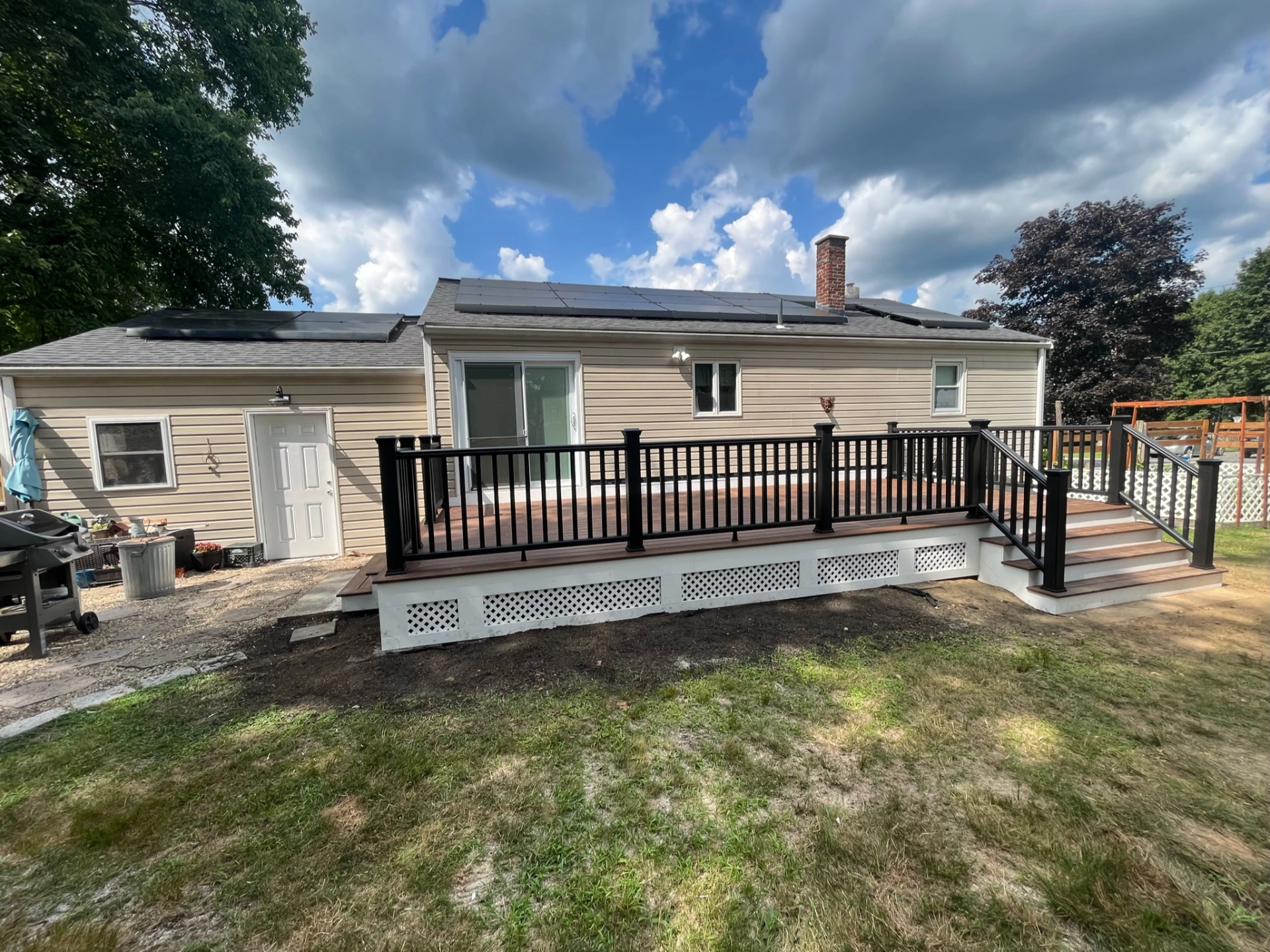 Backyard view of house with solar panels, wooden deck, black railing, and sliding glass door under cloudy sky.