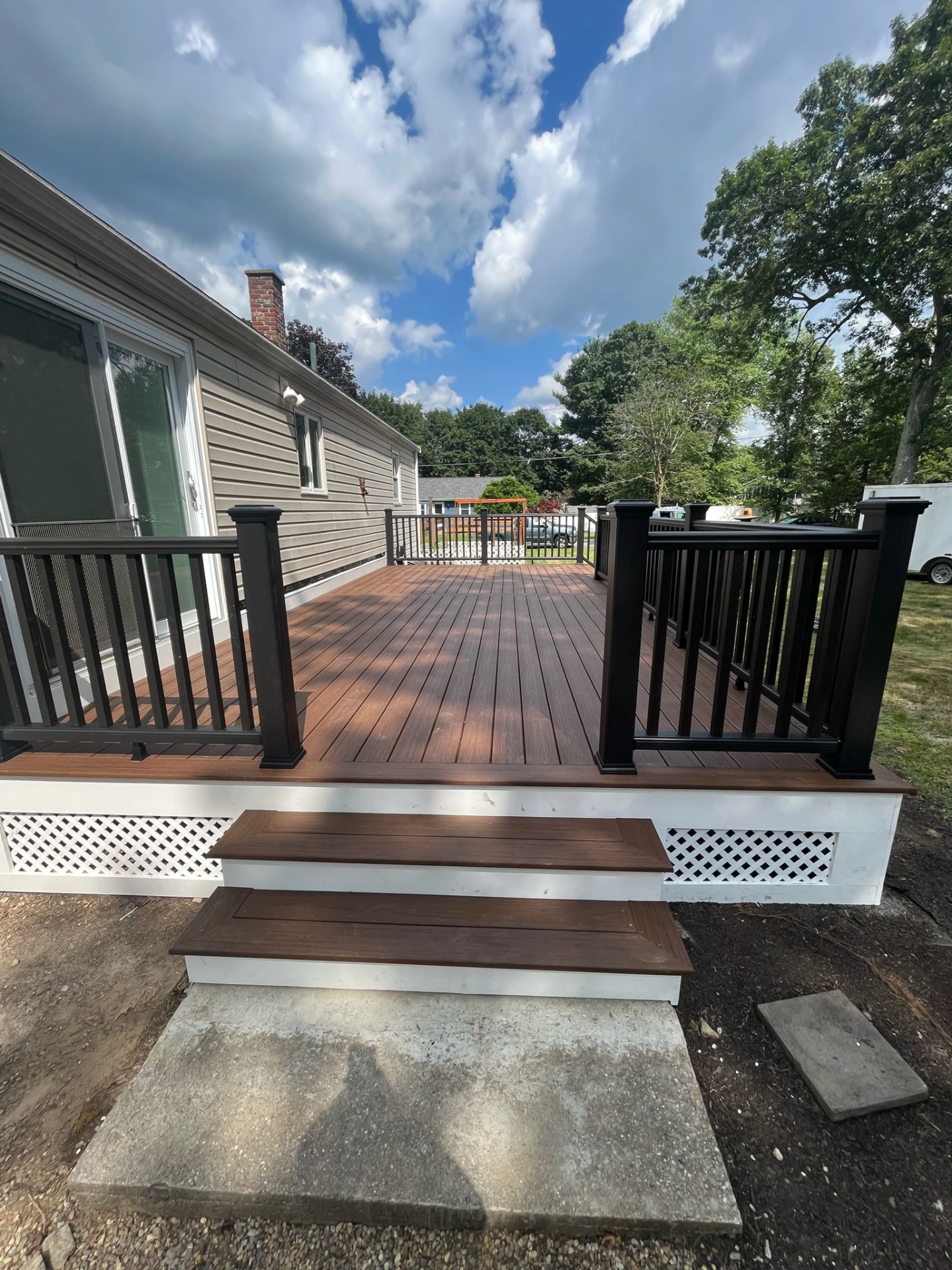 Wooden deck with black railing attached to beige house, steps leading down, surrounded by trees and cloudy sky.