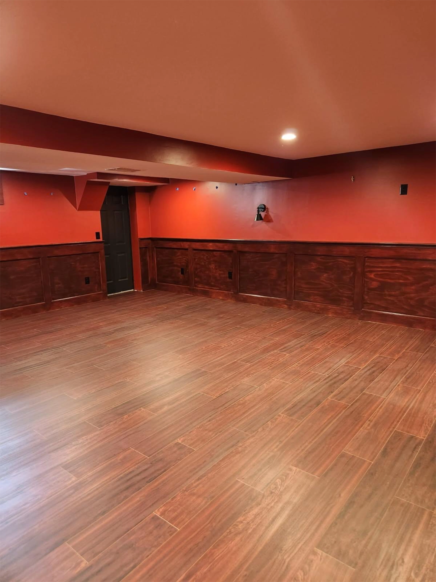 basement-room-with-brown-wood-paneling-and-flooring-red-walls-recessed-lighting-and-a-closed-black-door