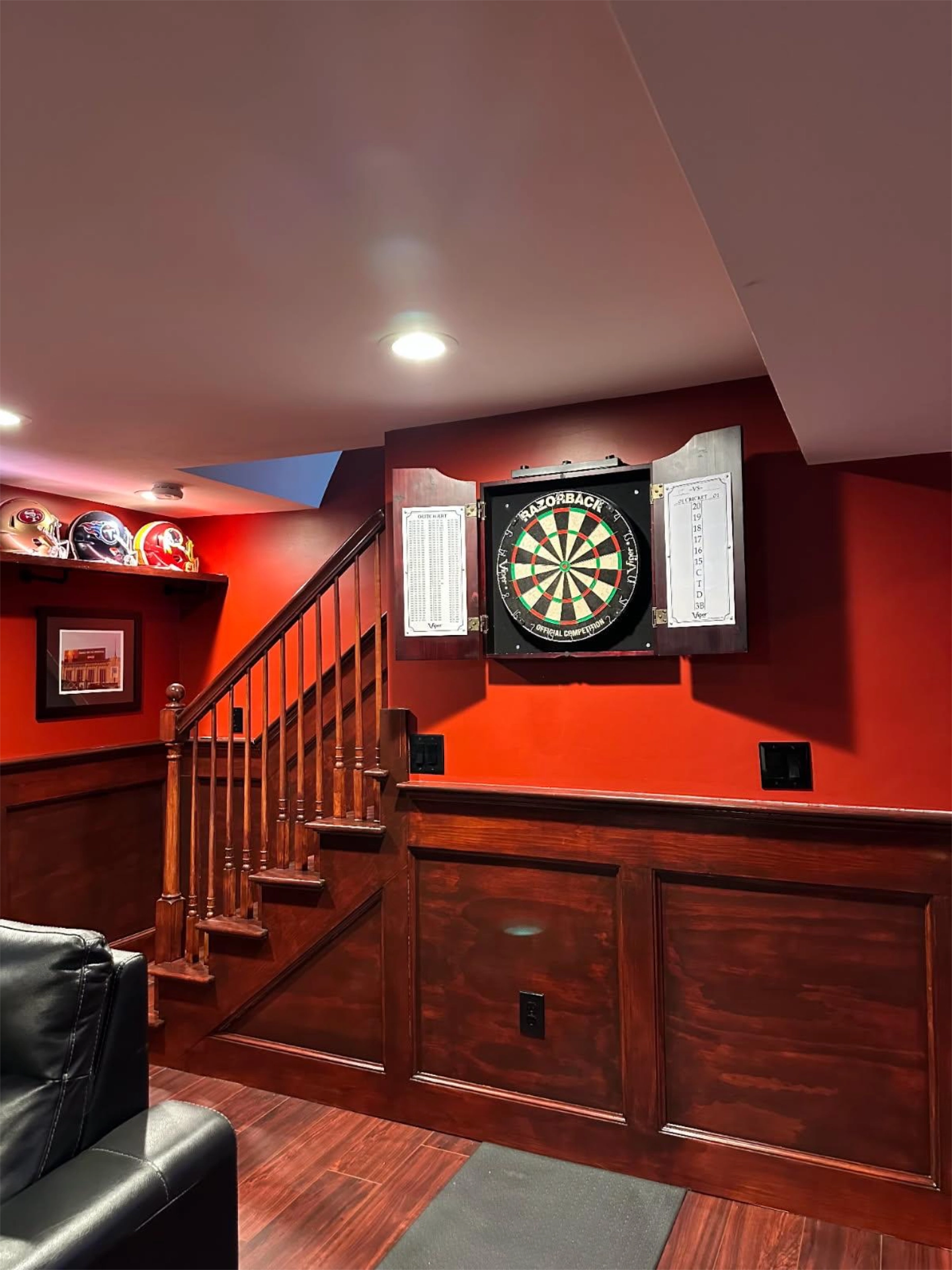dartboard-with-open-cabinet-doors-on-red-wall,-wooden-stairs-framed-picture-and-footbal-helmets-on-shelf-nearby-on-basement-in-needham-ma