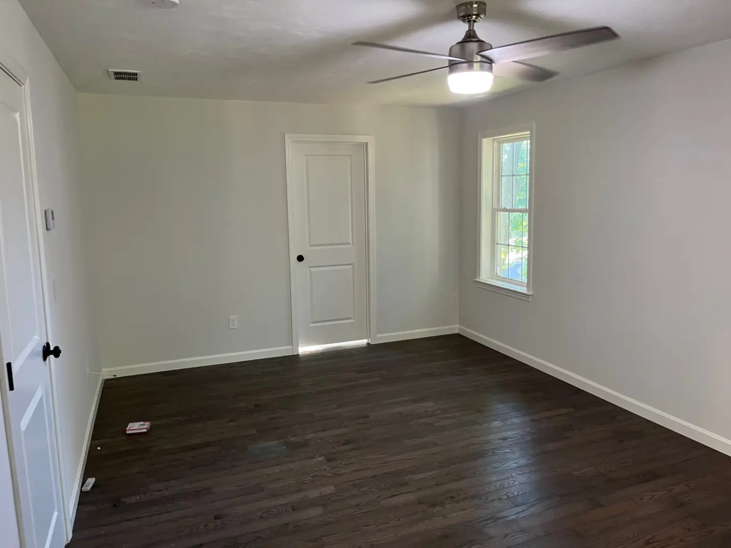 empty-room-with-white-walls-dark-wood-floor-ceiling-fan-window-with-view-of-trees-and-two-closed-white-doors-in-needham-ma