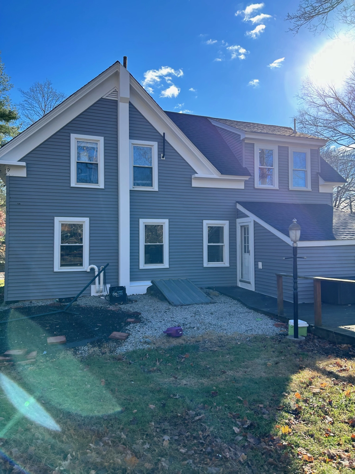 gray-two-story-house-with-white-trim-gravel-pathway-visible-sun-glare-and-surrounding-trees-in-needham-ma
