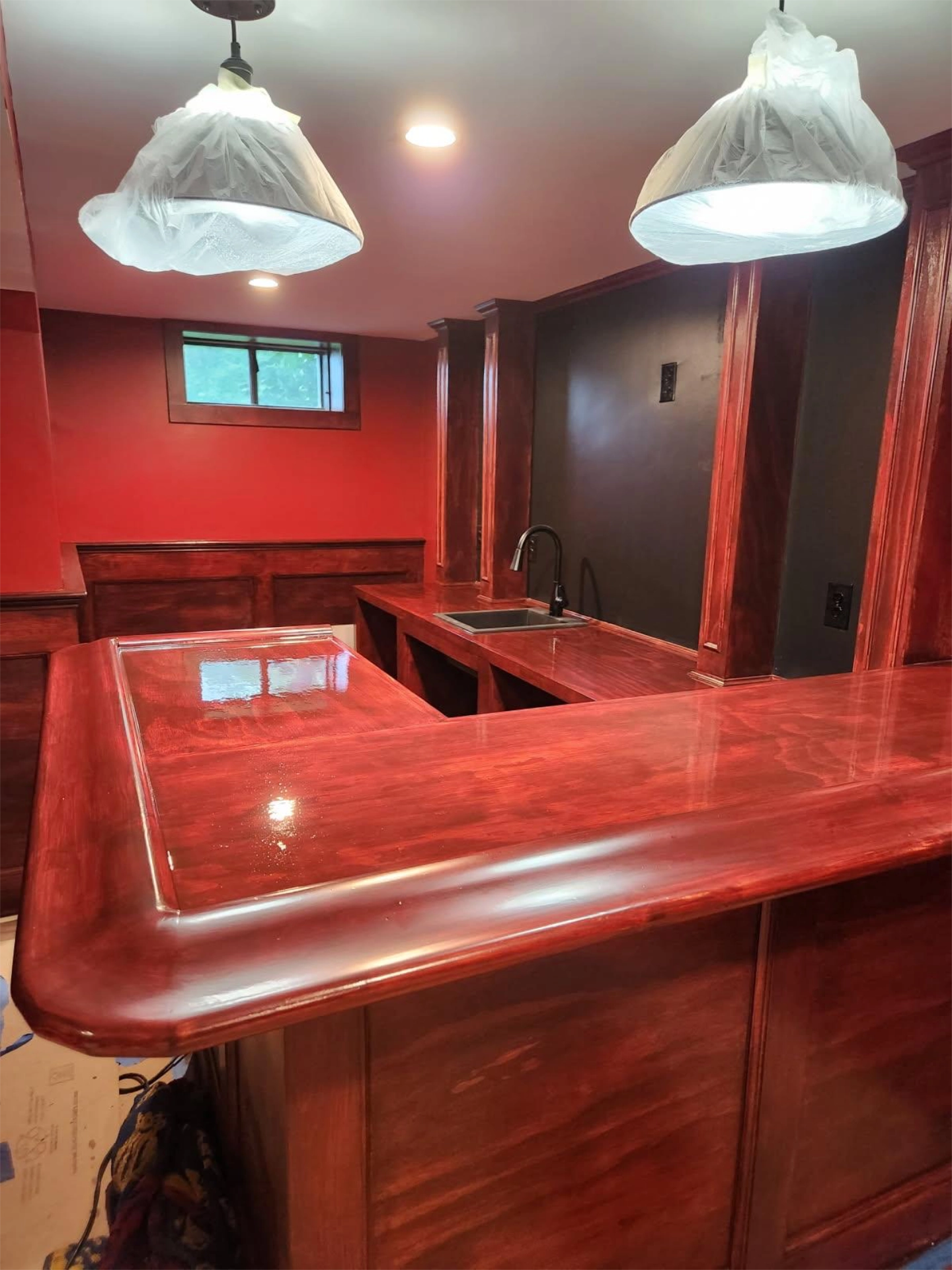 Wooden bar counter with glossy finish, overhead lights wrapped in plastic, red walls, small window, sink, and faucet.