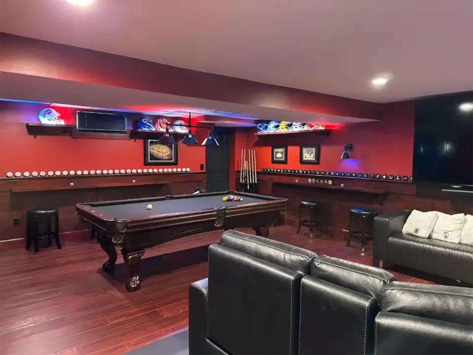 Basement game room with pool table, red walls, sports helmets on shelves, numerous baseballs, black stools, and a large TV.