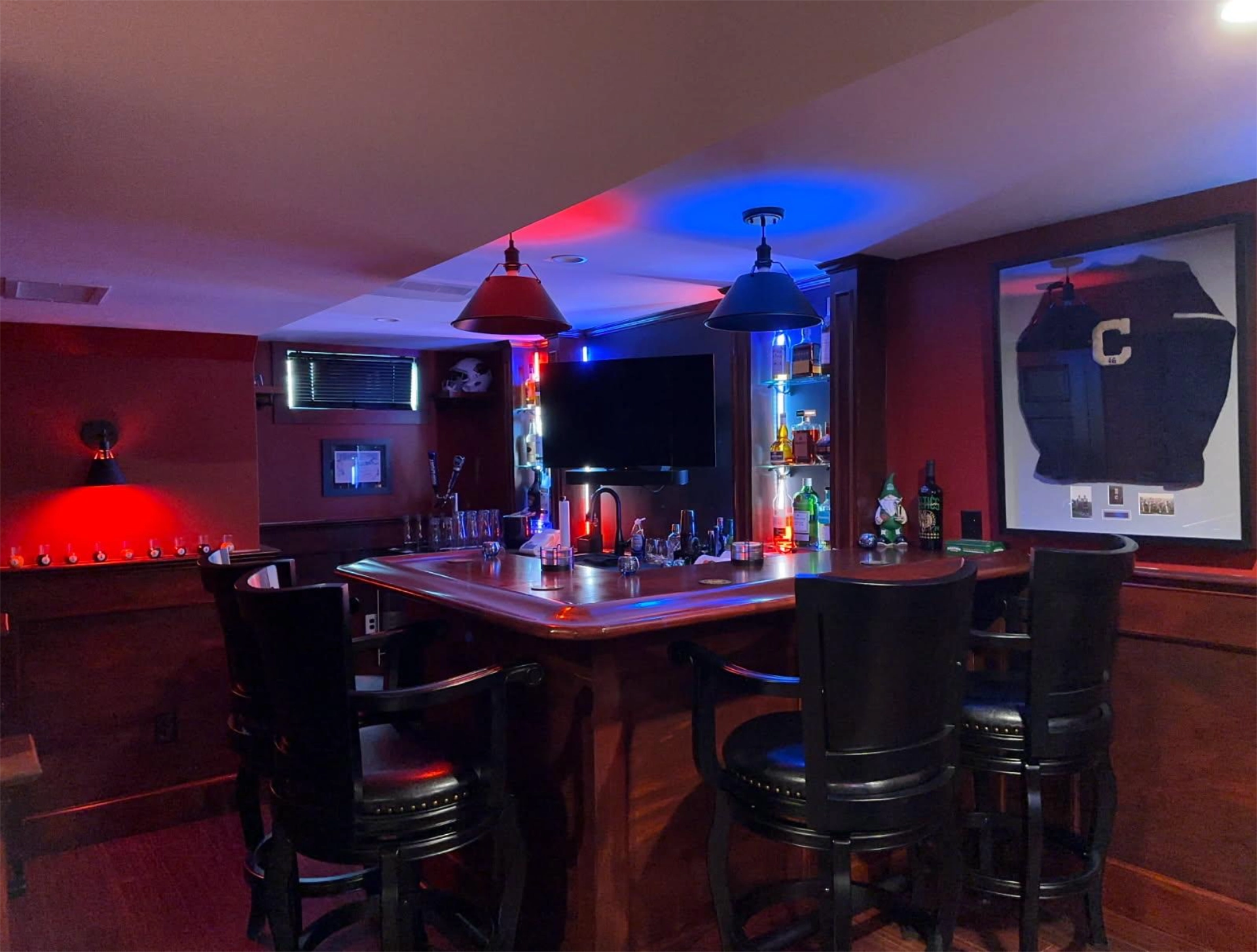 Home bar with curved counter, black chairs, framed jersey, red and blue lights, TV, liquor bottles on shelves.