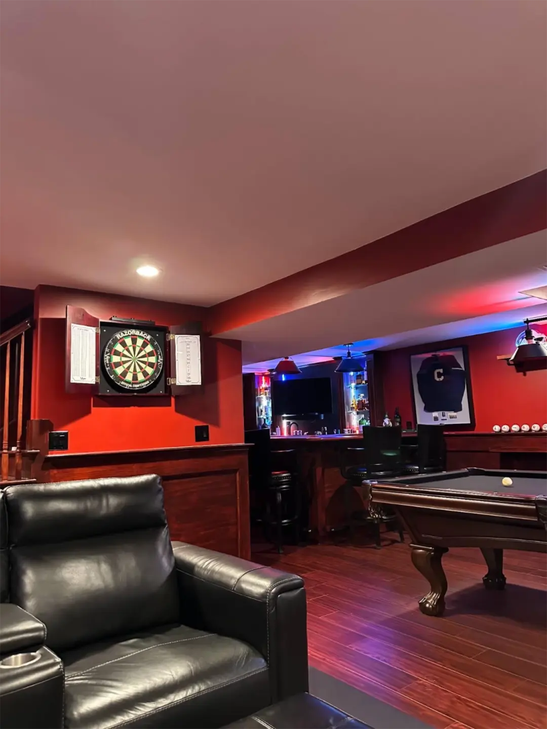 things-on-basement-is-dartboard-on-red-wall-above-black-leather-chair-pool-to-the-right-bar-area-with-chairs-and-sports-jersey-in-needham-ma